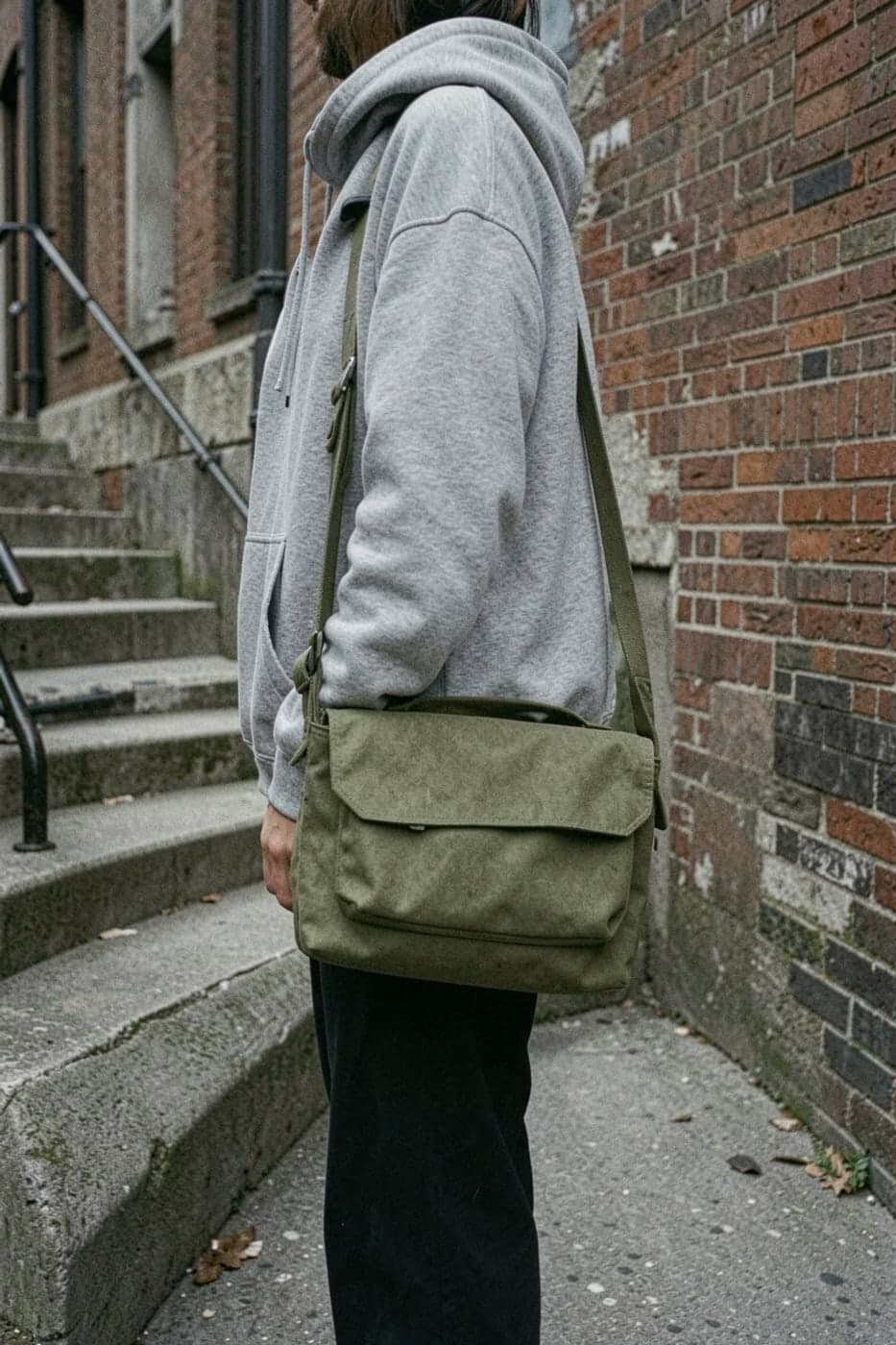 An olive canvas messenger bag worn over a grey hoodie on a city street